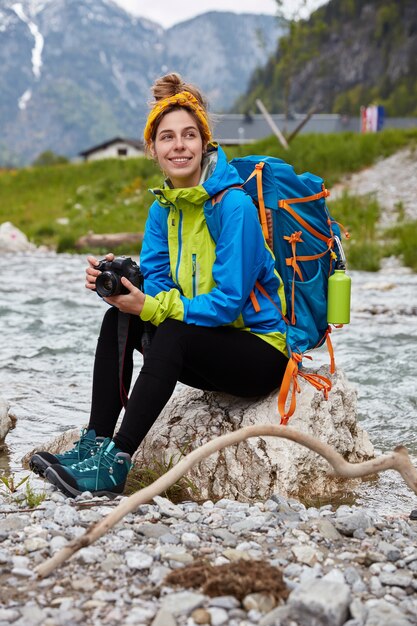 Thoughtful pleased tourist hiker takes part in adventure, has active rest in mountains, poses on stone near stream