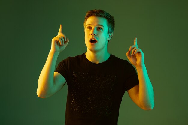 Thoughtful pointing up. Caucasian man's portrait isolated on green studio background in neon light. Beautiful male model in black shirt. Concept of human emotions, facial expression, sales, ad.