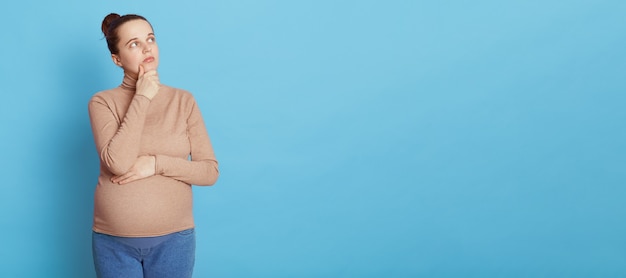 Free photo thoughtful pregnant woman looks thoughtfully aside, keeping hand on chin, makes planning about child birth, dreams to become mother, wearing beige sweater and jeans, isolated on blue wall.