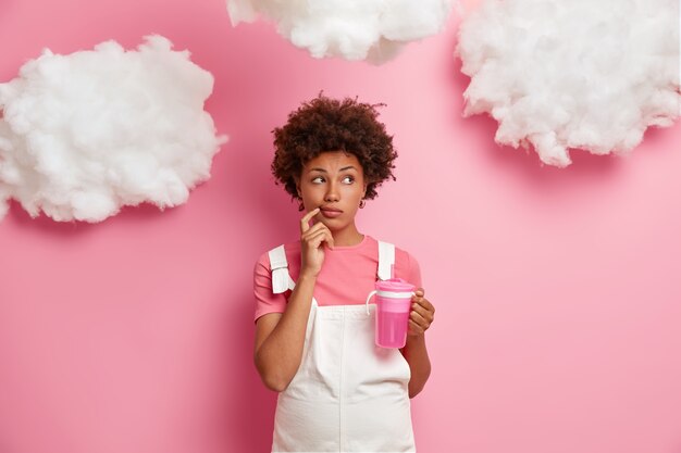 Thoughtful pregnant woman looks thoughtfully aside, makes planning about child birth, dreams to become mother, dressed in clothes for future moms, drinks water isolated on pink wall white clouds