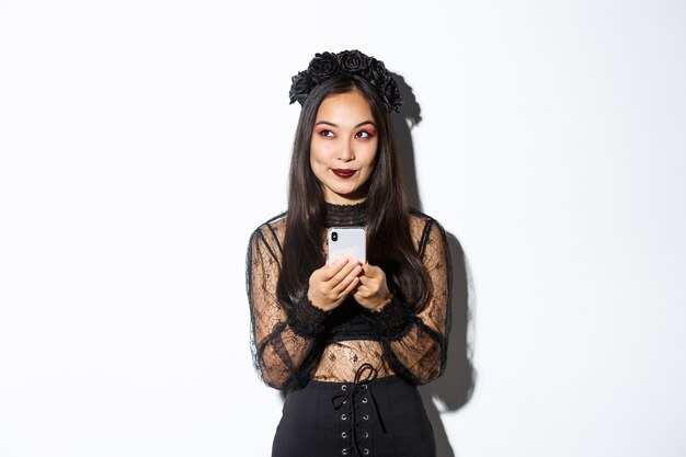 Free photo thoughtful pretty asian girl in gothic elegant dress holding smartphone while thinking