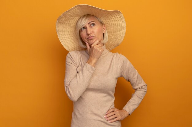 ビーチ帽子をかぶった思いやりのあるきれいな金髪のスラブ女性は、オレンジ色の側を見ているあごを保持します