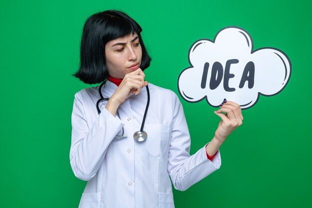 Thoughtful pretty caucasian girl in doctor uniform with stethoscope holding and looking at idea bubble