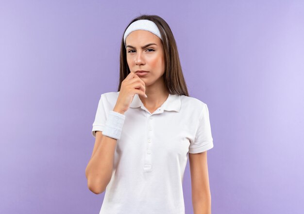 Thoughtful pretty sporty girl wearing headband and wristband putting hand on chin isolated on purple space