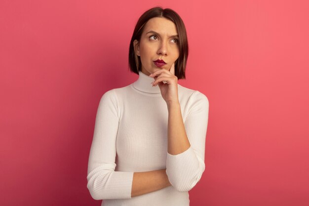 Thoughtful pretty woman puts hand on chin and looks at side isolated on pink wall