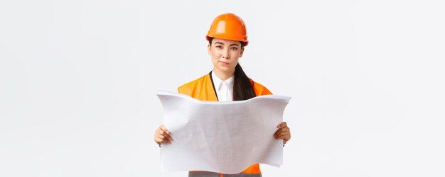 Thoughtful professional asian female construction manager architect reading blueprints and looking at camera pondering making decision while studying project plan white background