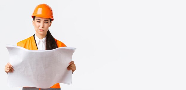 Thoughtful professional asian female construction manager architect reading blueprints and looking at camera pondering making decision while studying project plan white background