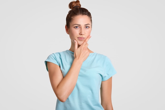 Thoughtful puzzled woman with hesitant expression, holds chin, dressed in casual clothes, ponders on something