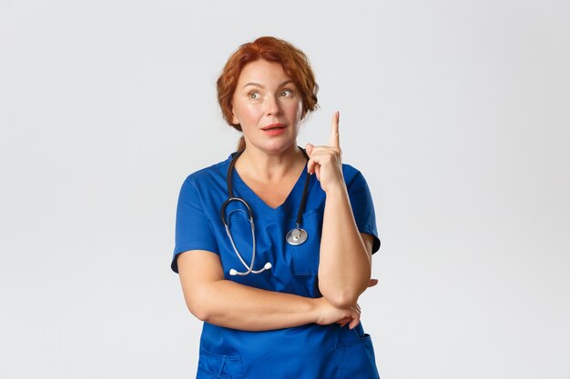 Thoughtful redhead middle-aged nurse, doctor in scrubs have assumption or suggestion, raising finger, have idea, share her thought, 