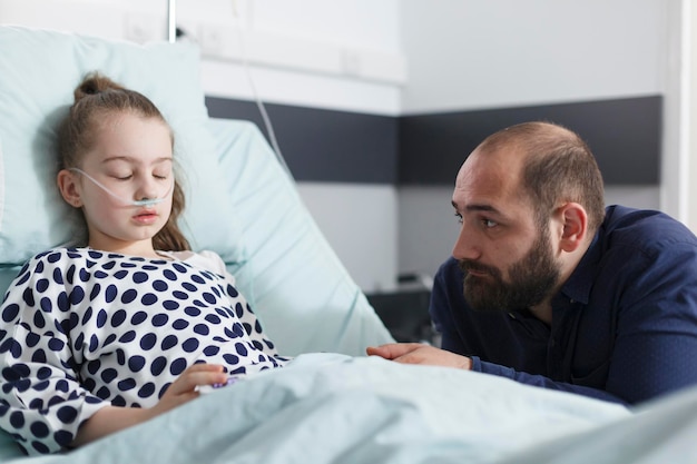 Thoughtful sad father looking at sleeping sick little daughter while in pediatric clinic patient room. Ill little girl resting after medical procedure while upset worried father sitting besides her.