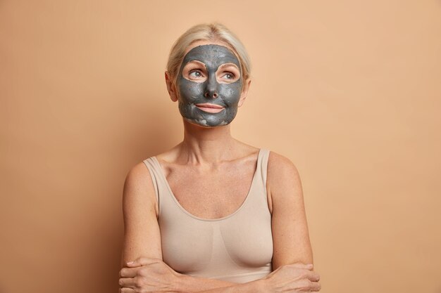 thoughtful senior blonde woman applies black choral beauty anti aging mask on face keeps arms folded dressed in casual top isolated on beige wall