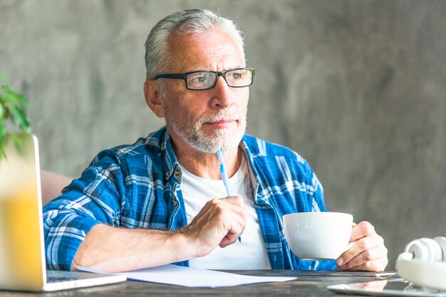 Thoughtful senior man holding pen and coffee cup making note