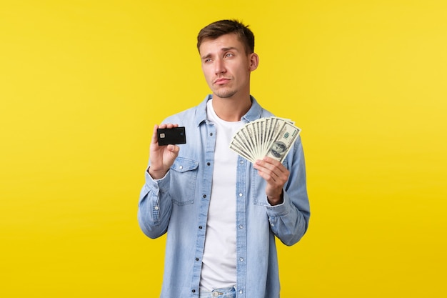 Thoughtful serious blond handsome man showing credit card and money, looking away thinking how invest money, pondering what buy during shopping, standing yellow background.
