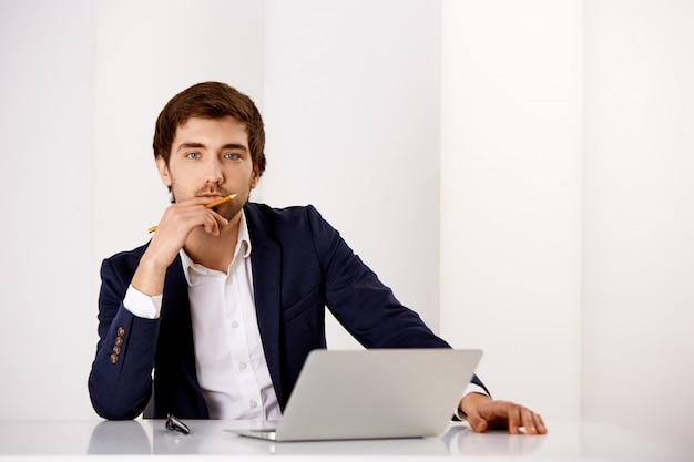 L'uomo intelligente premuroso in vestito si siede nel suo ufficio con il computer portatile, tocca il labbro come meditando, prendendo una decisione importante