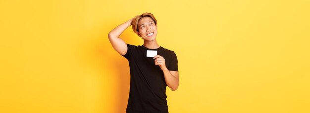 Thoughtful smiling asian guy thinking while showing credit card looking upper left corner dreamy yel