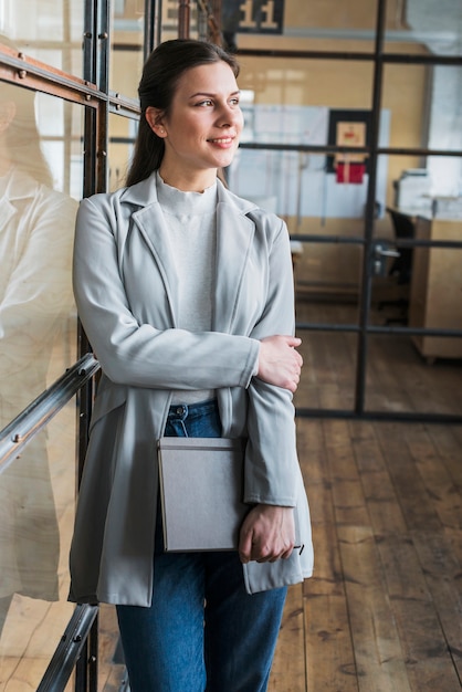 Thoughtful smiling businesswoman leaning on glass wall and holding diary in office