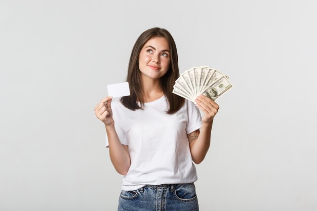 Задумчивая улыбающаяся девушка держит деньги и кредитную карту, смотрит в левый верхний угол, стоит белая и размышляет.