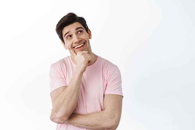 Thoughtful smiling man looks aside at logo with dreamy smile, imaging something interesting, have idea, standing and pondering against white background