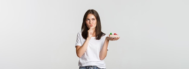 Free photo thoughtful smiling pretty girl pondering while holding piece of cake white background