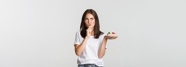 Thoughtful smiling pretty girl pondering while holding piece of cake white background