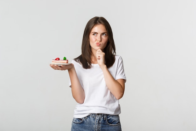 Thoughtful smiling pretty girl pondering while holding piece of cake, white.