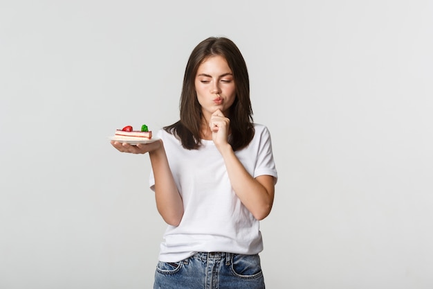 Thoughtful smiling pretty girl pondering while holding piece of cake, white.