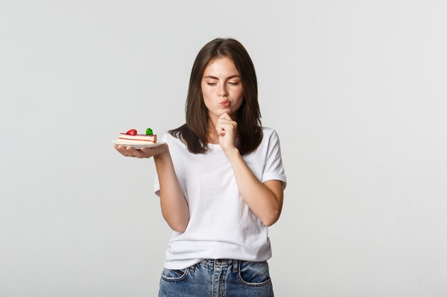 Thoughtful smiling pretty girl pondering while holding piece of cake, white.