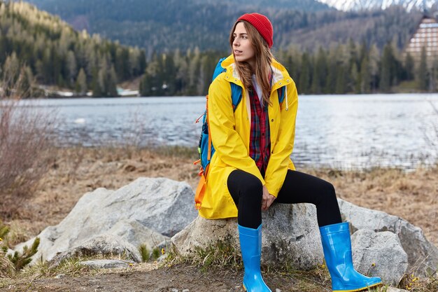 Thoughtful smiling tourist girl wears yellow raincoat and rubber boots sits on stone