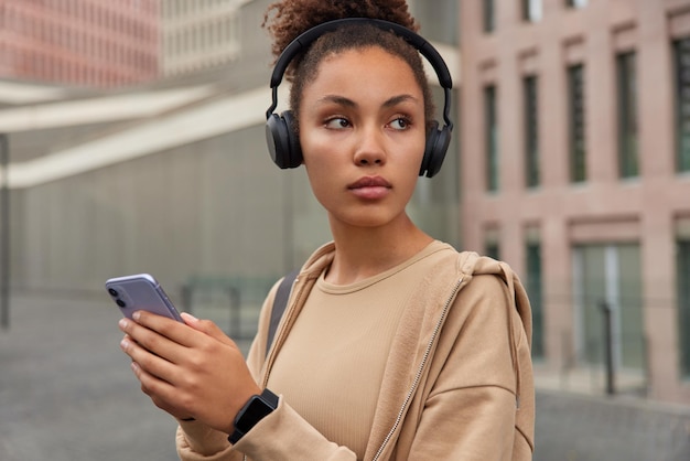 Thoughtful sporty woman walks outdoors listens motivational music for training wears wireless headphones on ears uses smartphone