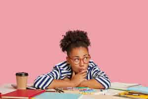 Free photo thoughtful student girl posing at the desk against the pink wall