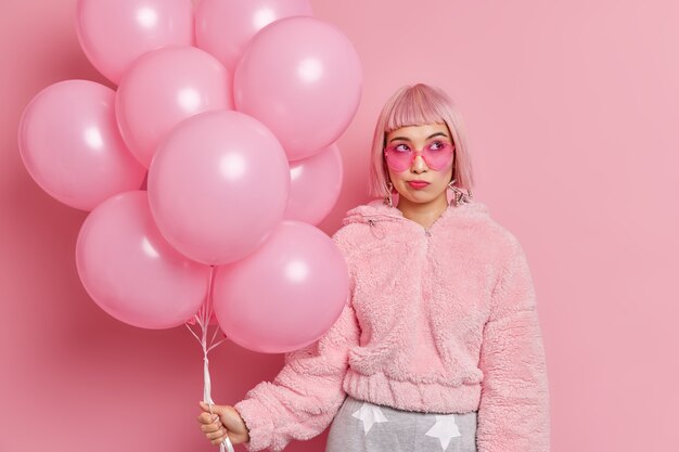 Thoughtful stylish young Asian woman has glamour look wears trendy sunglasses fur coat holds inflated balloons thinks what surprise to prepare for congratulating friend with special occasion.