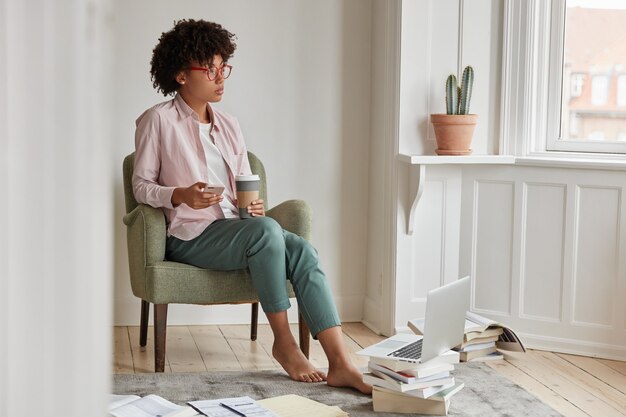 Thoughtful successful female startupper enjoys cappuccino beverage, holds takeaway cup, sits in armchair, uses cell phone and laptop computer