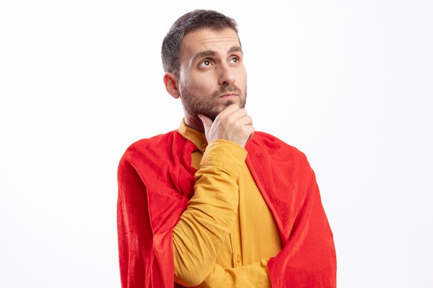 Thoughtful superhero man with red cloak puts hand on chin and looks at side isolated on white wall