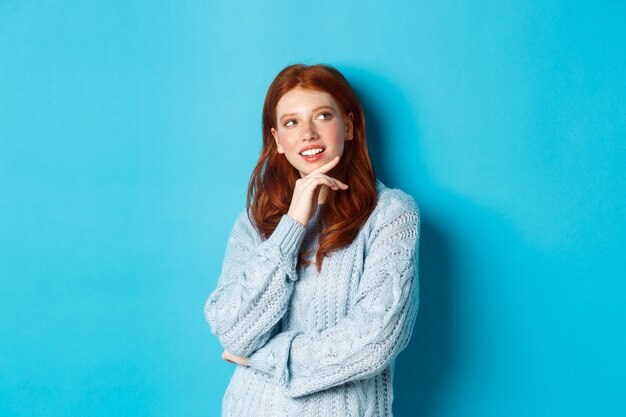 Thoughtful teenage girl with red hair, looking upper right corner logo and thinking, imaging something, standing over blue background