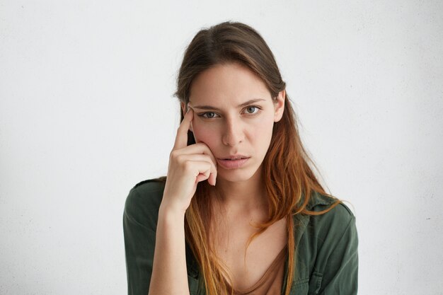 Thoughtful tired female having warm dark eyes and straight hair holding her index finger on temple looking seriously