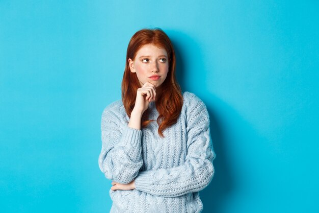 Thoughtful and upset redhead girl looking right, pondering solution, standing in sweater against blue background.