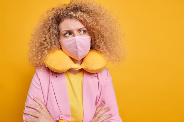 Thoughtful woman being on self isolation wears protective mask against coronavirus wears neck pillow keeps arms folded contemplates about something isolated over yellow wall with blank space