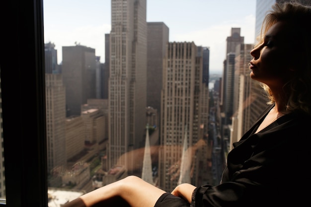 Free photo thoughtful woman in black silk robe sits on windowsill before beautiful new york cityscape