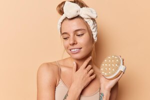 Thoughtful woman concentrated down touches face gently holds dry brush for exfoliating skin wears headband and t shirt isolated over beige background. cellulite treatment and dry brushing concept