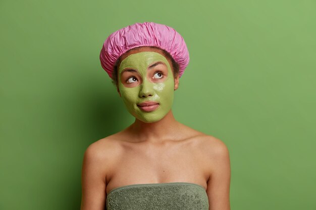 Thoughtful woman concentrated above wears green facial mask on face for rejuvenation wears bath hat towel around body thinks about how look beautiful enjoys skin care treatments