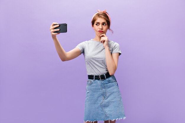 デニムスカートの思いやりのある女性は、紫色の背景で自分撮りを取ります。灰色のTシャツにピンクのバンダナを持つ少女が写真を作ります。