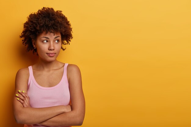 thoughtful woman has healthy skin, natural curly hair, keeps arms folded and looks pensively aside, wears casual clothing, isolated over yellow wall, tries to make decision