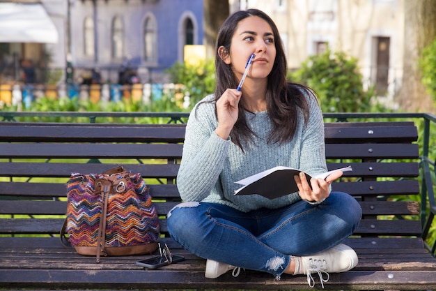 Thoughtful woman making notes and sitting on bench outdoors