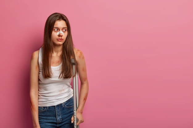 Thoughtful woman patient looks away, has bruised skin, doesnt have abilities to walk after accident, poses with crutches. Female slipped on winter road, has various injuries. Medical concept