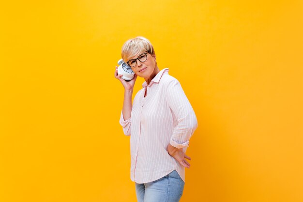 Thoughtful woman in pink shirt listens to alarm clock on orange background