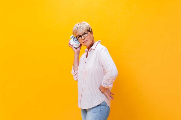 Thoughtful woman in pink shirt listens to alarm clock on orange background