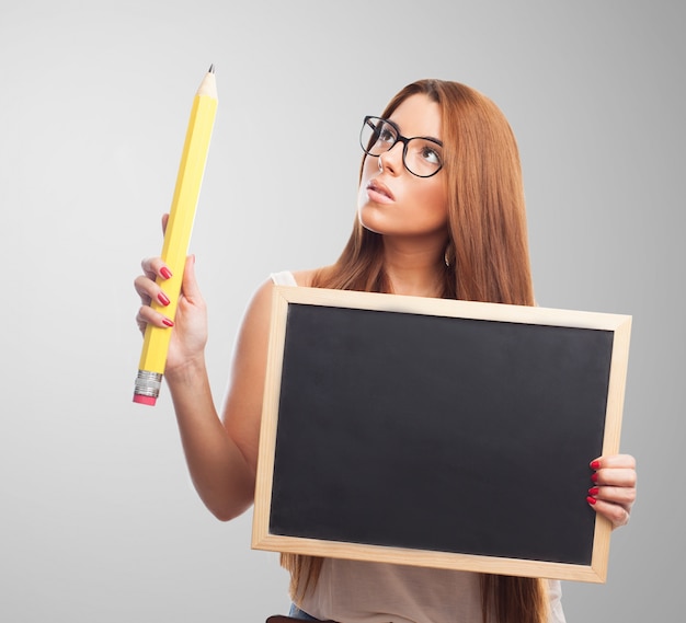 Thoughtful woman in spectacles holding blackboard and big pencil