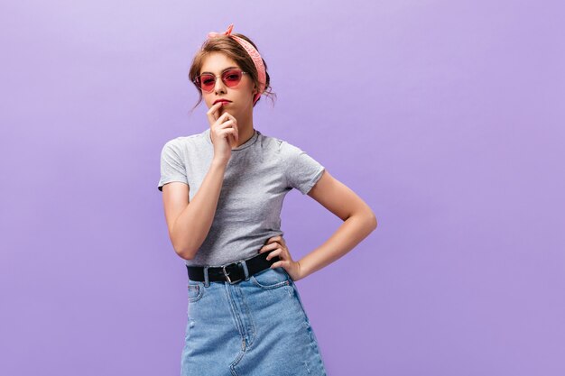 Thoughtful woman in sunglasses and grey shirt poses on purple background. Pretty young lady in pink headband and summer clothes looking into camera.