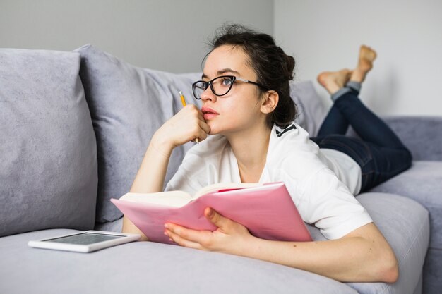 Thoughtful woman with book lying on couch