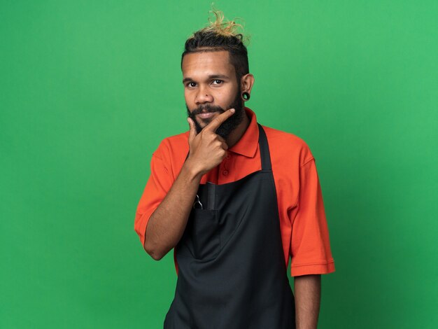 Thoughtful young afro-american male barber wearing uniform keeping hand on chin  isolated on green wall with copy space